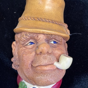 May include: A detailed ceramic figurine of a man's head, wearing a tan hat and smoking a white pipe. The figurine has blue eyes, a red collar, and a green and blue emblem. The face is detailed with wrinkles and a smiling expression.