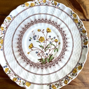 May include: A white ceramic plate with a scalloped edge and a basket-weave texture. The center features a botanical illustration of yellow buttercups and small blue berries. A brown decorative border surrounds the central floral design.