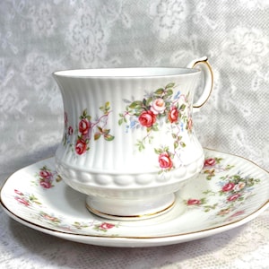 May include: White teacup and saucer set with a floral rose pattern. The teacup has a ribbed design and a gold handle. The saucer and teacup are decorated with red and pink roses, green leaves, and gold trim.