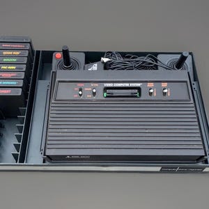 May include: A black Atari 2600 Video Computer System console housed in a plastic organizer tray. The set includes two black joysticks with red buttons, a power cable, and seven game cartridges labeled Football, Home Run, Berserk, Pac-Man, Swordquest, Space Invaders, and Combat.