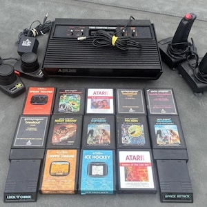 May include: A black Atari 2600 video game console with two black joysticks, two paddle controllers, and thirteen game cartridges including titles like Pac-Man, Defender, Breakout, Space Invaders, and Raiders of the Lost Ark.