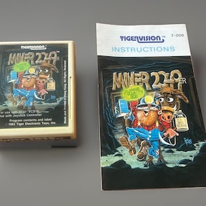 May include: A beige Atari VCS video game cartridge and its matching instruction manual for the game Miner 2049er by Tigervision. Both items feature a colorful illustration of a bearded miner character and a horse in a dark mine setting.