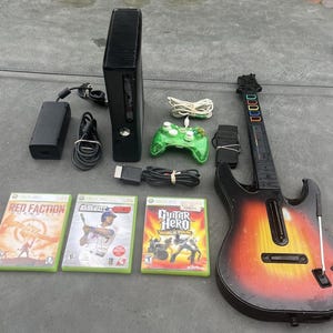 May include: An Xbox 360 console with a green controller, a Guitar Hero guitar, and three game cases: Red Faction, Baseball 2K8, and Guitar Hero World Tour. The console and accessories are black.