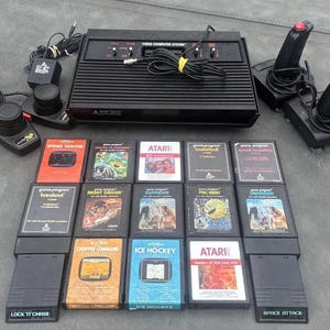Puede incluir: Una consola de videojuegos Atari 2600 negra con dos joysticks negros, dos controladores de paleta y trece cartuchos de juego, incluyendo títulos como Pac-Man, Defender, Breakout, Space Invaders, Night Driver y Raiders of the Lost Ark.