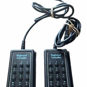 May include: Two black rectangular keyboard controllers with attached black cables. Each device features a numeric keypad with buttons labeled 1 through 9, 0, an asterisk, and a pound sign. The text Keyboard Controller is printed in light blue.