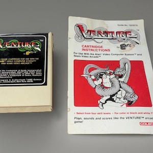 Puede incluir: Un cartucho de videojuego Atari Venture y una hoja de instrucciones. El cartucho está en una caja beige con una etiqueta negra. La hoja de instrucciones presenta un fondo rojo con una ilustración gris.