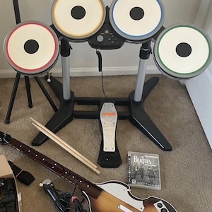 May include: A video game drum set with four white pads featuring red, yellow, blue, and green rims, a black foot pedal, two wooden drumsticks, a wired microphone, and a guitar controller with a sunburst finish and The Beatles Rock Band game case.
