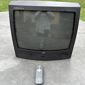Puede incluir: Un televisor de tubo de rayos catódicos RCA XL100 Commercial Skip negro con pantalla de cristal curvada y un mando a distancia negro a juego con teclado numérico, botones direccionales y varios controles de función situado frente a la unidad.