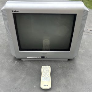 May include: A silver RCA TruFlat cathode ray tube television with a dark screen and a matching off-white remote control. The television features a rectangular frame with integrated side speakers and a front-facing control panel below the screen.