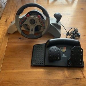 Op de afbeelding: Een zwart en grijs PlayStation 2-racewiel met een zilveren basis en een zwarte pedaalset. Het stuur heeft een rood en zwart ontwerp met een PlayStation 2-logo op de pedaalset.