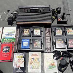 May include: A vintage Atari Video Computer System console with wood grain trim, two black joystick controllers, two paddle controllers, and eight game cartridges including Missile Command and Fishing Derby. Includes several instruction manuals.