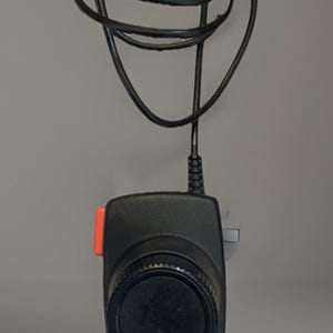 May include: Black electronic device with a dial and an orange button. The device has a black cord and the word "driving" with a car graphic is printed on the front. The cord is looped and secured with a green tie.