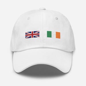 May include: A white baseball cap featuring two small rectangular flag icons embroidered on the front. The left icon displays the Union Jack flag, while the right icon displays the vertical green, white, and orange tricolor flag of Ireland.