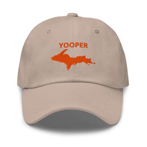 Può includere: Un cappellino da baseball beige con un ricamo arancione della penisola superiore del Michigan e il testo YOOPER centrato sopra la mappa. Il cappellino presenta una visiera curva, una corona strutturata e occhielli di ventilazione.