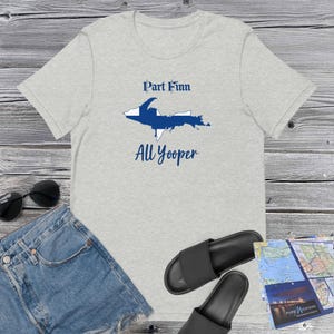 Peut inclure: Un t-shirt gris clair avec le texte "Part Finn All Yooper" en bleu marine. Le motif comprend un contour bleu et blanc d'un état. Le t-shirt est présenté avec un short en jean, des lunettes de soleil, des sandales et une carte.