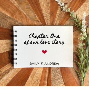 May include: A spiral-bound notebook with the text "Chapter One of our love story" and a red heart. The names "EMILY & ANDREW" are printed below. The notebook is on a wooden surface with a floral accent.