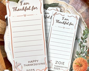 May include: White and brown Thanksgiving gratitude cards with the text "I am Thankful for" and lines for writing. The cards have a decorative border and are tied with a green ribbon. The words "Happy Thanksgiving 2025" are also printed on the cards.