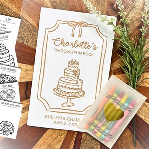 May include: A white wedding activity book with gold foil text reading Charlotte's Wedding Fun Book and Evelyn & Charlotte June 2, 2026, featuring a three-tier cake illustration, alongside a translucent pouch of colorful crayons with a gold heart sticker.