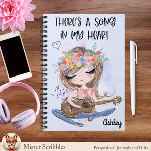 May include: Spiral-bound notebook with the text "THERE'S A SONG IN MY HEART" and an illustration of a girl playing a guitar. The girl is wearing a floral crown and jeans. A white pen, headphones, and a smartphone are on the wooden surface.