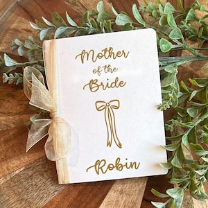May include: A white card with gold text that reads "Mother of the Bride Robin" with a bow illustration. The card is tied with a sheer, gold ribbon. Greenery and a wooden surface are in the background.