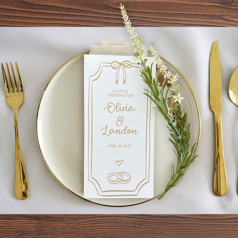K&ouml;nnte beinhalten: Eine Hochzeitstafel mit einem wei&szlig;en Teller, goldenem Besteck und einer personalisierten Karte. Auf der Karte steht "A Little Wedding Fun" und die Namen "Olivia & Landon" sowie das Datum "25. April 2027". Ein Zweig Gr&uuml;nzeugs sorgt f&uuml;r einen nat&uuml;rlichen Touch.