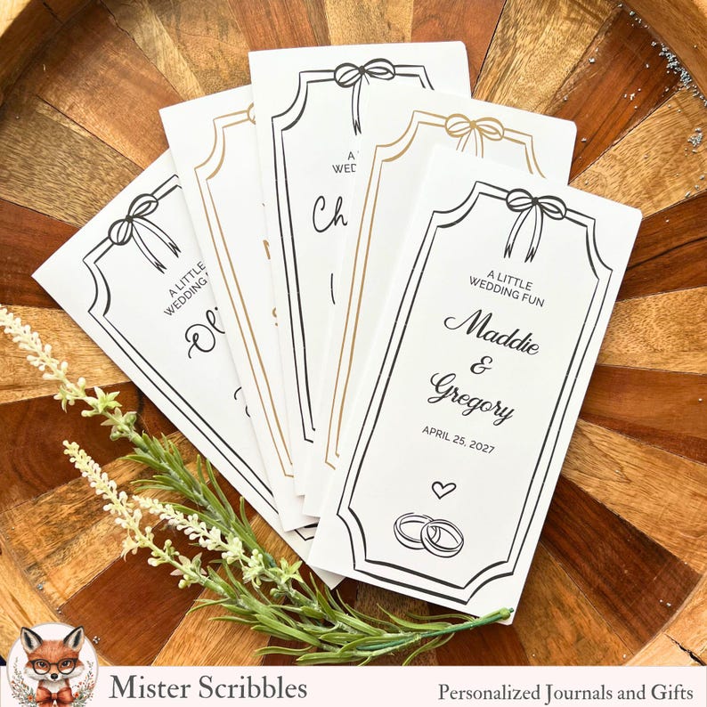 K&ouml;nnte beinhalten: Eine Reihe wei&szlig;er Hochzeitskarten mit schwarzen und goldenen Akzenten. Jede Karte hat ein Schleifendesign und die Worte "A Little Wedding Fun". Eine Karte ist personalisiert mit den Namen "Maddie & Gregory" und dem Datum "April 25, 2027."