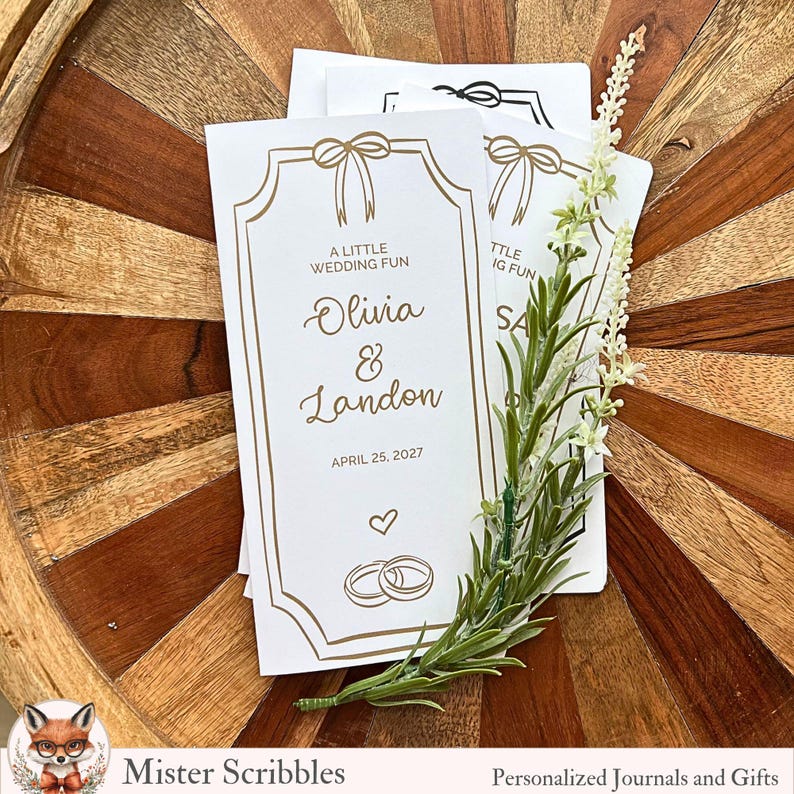 K&ouml;nnte beinhalten: Wei&szlig;e Hochzeitskarten mit goldenen Akzenten, mit den Namen Olivia & Landon und dem Datum 25. April 2027. Die Karten sind mit Schleifen- und Ringgrafiken verziert. Ein Zweig Gr&uuml;n liegt auf den Karten, die sich auf einer Holzoberfl&auml;che befinden.