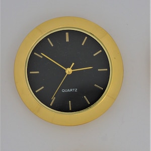 May include: Gold-toned clock face with black hands and black numerals. The clock face is labeled "QUARTZ" in white lettering.