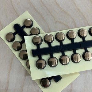 May include: Close-up of two sheets of gold-colored, round-headed rivets attached to black plastic strips. The rivets are on a light yellow adhesive backing. The number "60065" is printed on one of the strips.