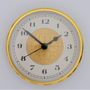 May include: A gold-rimmed clock face with black hands and white Roman numeral hour markers. The clock face has a decorative, gold-colored pattern in the center.