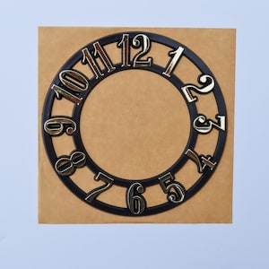 May include: A black clock face with metallic gold numerals, arranged in a circular pattern. The numbers are set against a brown square background, with a light blue backdrop.