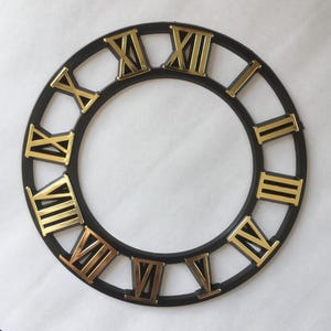 May include: A black clock face with gold Roman numerals. The circular frame is black, and the numerals are in a gold color. The clock face has a classic, elegant design, suitable for home decor.