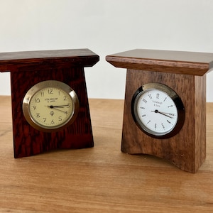 May include: Two wooden clocks with a simple design. The clocks have a square base with a small shelf on top. The clock faces are round and have black Roman numeral markings. One clock face is white with black hands and the other is cream with gold hands. The clock on the left has the text "SCHLAGE & SONS" on the face.