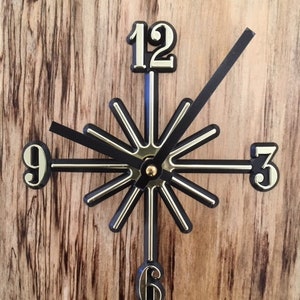 May include: A black and gold clock with a unique design. The clock face has a starburst pattern with black hands and gold accents. The numbers 12, 9, 6, and 3 are displayed on the clock face.