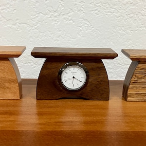 May include: Three wooden clock stands with different wood finishes. Each stand has a small round clock face with black hands and Roman numeral markings.