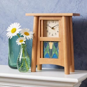 May include: A wooden mantel clock with a square face and a decorative tile insert. The clock has a light wood finish and a cream-colored clock face with black numerals. A vase of daisies sits to the left of the clock.