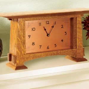 May include: A wooden clock with a copper face and black Roman numeral markings. The clock is sitting on a white shelf.