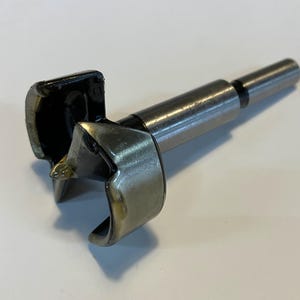 May include: A silver and black drill bit with a pointed tip and a black plastic collar. The drill bit is designed for creating holes in wood.