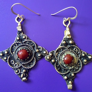 May include: A pair of silver earrings with intricate designs and red gemstone accents. The earrings are shaped like a square with rounded corners and feature a central red stone.