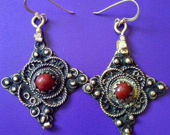 May include: A pair of silver earrings with intricate designs and red gemstone accents. The earrings are shaped like a square with rounded corners and feature a central red stone.