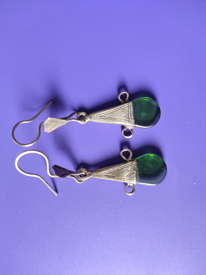 May include: A pair of silver earrings with green glass teardrop-shaped stones. The earrings have a geometric design and a hook closure.