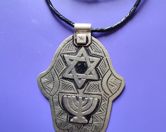 Pode incluir: Um colar com um pingente de prata em forma de mão de Hamsa com uma Estrela de David e um candelabro. O colar tem um cordão preto.