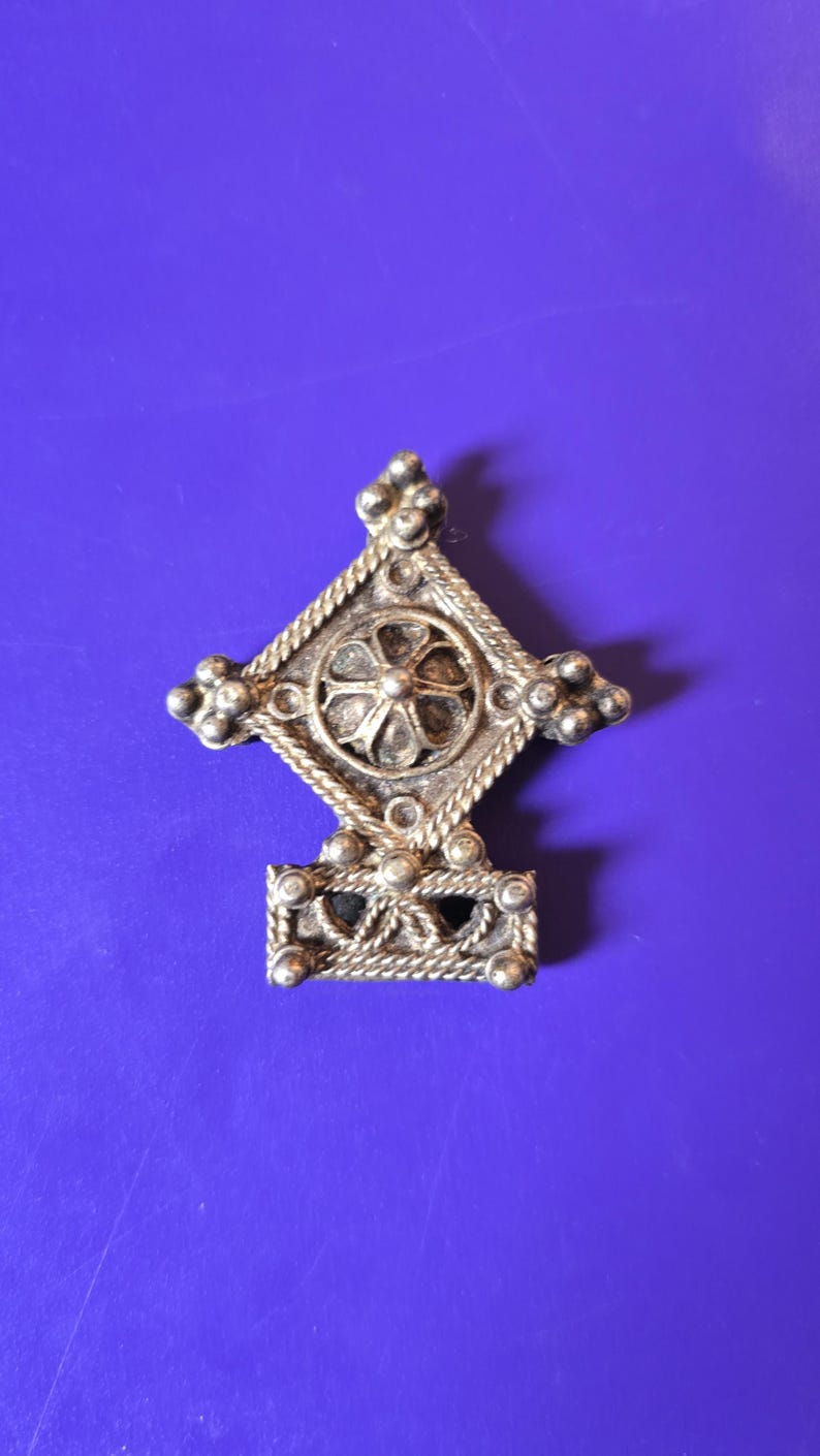 May include: A silver-toned metal brooch featuring a diamond-shaped upper section with a central circular floral motif, surrounded by twisted rope detailing and small spherical accents, with a rectangular base featuring a braided pattern.