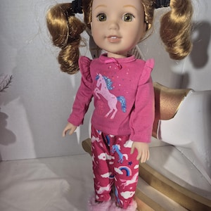 May include: A doll with auburn hair styled in pigtails, wearing a pink long-sleeved shirt with a unicorn graphic and matching pajama pants. The pajama pants have a unicorn and rainbow pattern. The doll is wearing pink fuzzy slippers.