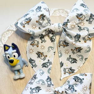 May include: Two white fabric hair bows with a repeating pattern of a cartoon dog, sun, and rainbow. The bows are tied together with a white ribbon.