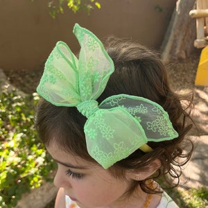 May include: A green sheer bow headband with floral embroidery. The headband is worn by a child with curly brown hair.