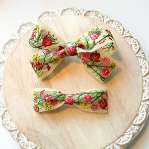 Op de afbeelding: Twee met bloemen geborduurde haarstrikken op een houten oppervlak. De grotere strik is crèmekleurig met rode, roze, gele en groene bloemen. De kleinere strik is crèmekleurig met roze, gele en groene bloemen.