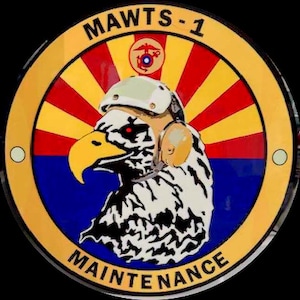 May include: Circular emblem with the text "MAWTS-1" and "MAINTENANCE". The emblem features a stylized eagle wearing a helmet, set against a red, yellow, and blue radial background. A smaller emblem is at the top.