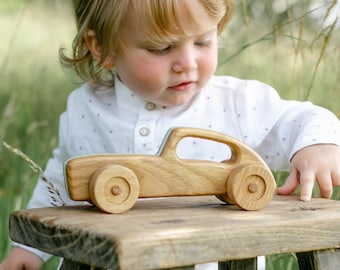 May include: A wooden toy car with a classic design. The car is made of light-colored wood and features round wheels. It sits on a rustic wooden stool, with a child nearby. The scene is set outdoors in a grassy area.