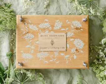 May include: A wooden box with a floral design and the text "BLUE FRONTIDE" is surrounded by greenery and flowers. The box is light brown with a white floral pattern and silver hardware. A pair of scissors is visible.